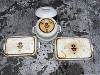Set of three white enamel cast iron cookware pieces seen from top down on a dark surface; includes lidded pot and two rectangular trays, all showing rust and wear marks, bottom of pieces visible