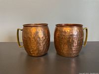 Two copper mugs shown side by side with hammered texture and brass handles under neutral lighting on a flat surface, viewed from the front with some visible patina and aging marks.