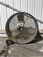 Front view of large industrial floor fan showing metal grille, blades, wheels, and power cord.