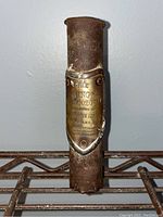 Full view of the rusted metal shield plaque with embossed text, mounted on a cylindrical metal base against a plain background.