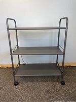Full front view of 3-tier metal rolling cart with flat gray shelves and caster wheels.