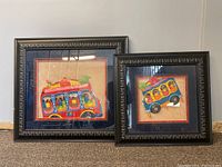 Frontal view of two framed South American textile art pieces leaning against a wall. Larger piece is on the left, smaller on the right. Frames are dark wood with carved patterns. Textile brightly colored with red, blue, yellow, green showing buses filled with people and items.