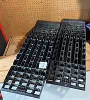Pair of black plastic car ramps with a grid-patterned design lying side by side on a wooden surface in front of a pegboard.