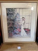 Framed art showing a woman in white feeding birds inside a large birdcage with flowers and plants around.
