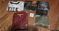 Photo of six folded clothing items including a white VICK sports jersey, maroon USC pullover, and four band/concert shirts mostly wrapped in plastic on a wooden floor.