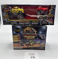 Yellow police rescue toy truck with red dirt bike on trailer in packaging along with two monster truck toy boxes.