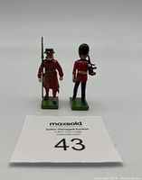 Two metal guard figures by W. Britains, each standing on green bases, front view showing detailed red uniforms and accessories.