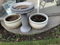 Photo showing a concrete bird bath placed between two white/cream ceramic pots with soil inside, set on concrete patio ground with grass in foreground.