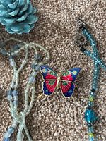 Photo showing a red, blue, green, and gold enamel butterfly pin alongside a multi-strand necklace with translucent and opaque light blue beads on carpet.