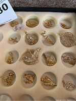 Full view of most of the gold electroplated charms displayed in a foam tray insert showing various shapes including hearts, crosses, animals, and decorative designs.
