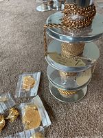 Four metal spools stacked, each wrapped with different thicknesses of gold-colored chain material, plus assorted small bags containing gold necklace adornments on carpet.