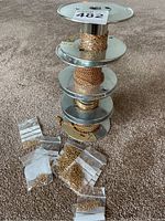 Four spools of gold tone chain arranged vertically with multiple small plastic bags of gold jump rings on the carpet beside them.