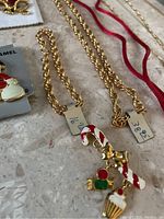Close-up image showing gold-tone chains with a candy cane pendant featuring red, white stripes, a bow, and holly berries embellishment. Also showing a Santa Claus enamel pin.
