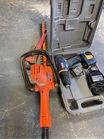 Photo showing Ryobi drill, battery, charger in plastic case, Black and Decker hedge trimmer, red pipe wrench, and sledgehammer on concrete floor.