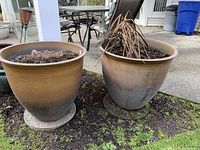 Two large brown outdoor plant pots placed on outdoor ground, each containing soil and dried plant remains, showing some dirt stains on pots from outdoor use.