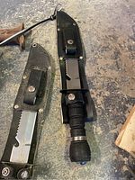 Two survival knives with black handles in leather sheaths on concrete floor