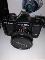 Front view of Yashica FX-3 Super 2000 camera with lens, showing focus and aperture rings, and lens cap with Yashica logo.
