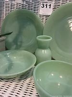 All 5 jadeite dinnerware pieces arranged on white wicker surface showing color and shape of large plate, small plate, two bowls and egg cup