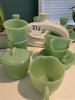 Photo showing multiple Fire King Jadeite items including creamer, sugar bowl with lid, cups, and saucers on a table with white decorative piece in background (not included).