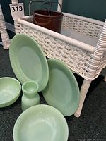 Photo of the jadeite green plates, bowls, egg cup arranged in front of a white wicker stand and brown clay container in background