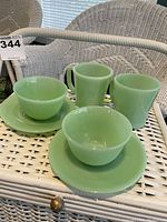 Photo 1 shows four pieces: two jade green jadeite tea cups on matching saucers and two mugs placed side by side on a white wicker chair.