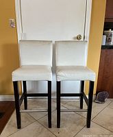 Front view of 2 wooden frame bar stools with white cushioned seats and backs, metal footrests.
