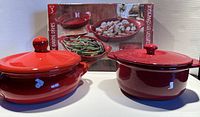 Two red ceramic baking dishes with lids in front of packaging box with images of dishes serving food