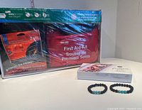 Photo showing sealed red first aid kit pouch with packaging indicating outdoor survival kit and easy access bandages, white box of essential oils, and two black lava bead bracelets with turquoise beads.