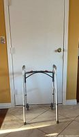 Metal folding walker with two front wheels and adjustable height standing against a white door on tiled floor.