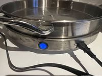 Close-up side view showing brushed stainless steel housing, blue illuminated power switch button, power cord, and base stand.