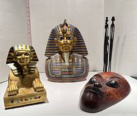Overview of all items including the golden Sphinx statue, King Tutankhamun bust statue, wooden mask, and family carving placed on a white surface with a measuring tape to show size.