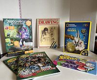 Photo showing all items included in the lot together on a white surface: National Geographic geode kit, Discovery crystal growing kit, drawing instruction book, two raised velvet coloring books, and a pack of scented markers.