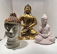 Front view of three Buddha statues: one large wooden gold-toned meditating Buddha in the center, medium sized white ceramic meditating Buddha with brown hair detail on right, and ceramic Buddha head on left.