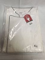Folded white chef coat in factory plastic packaging with tags visible. Buttons and black piping along collar shown.