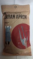 Factory packaged Chef Works Berkeley Urban Bib Apron in sky blue with green strap, photo of front packaging showing product name and illustration of apron.