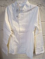 Full view of one white Chef Works chef coat hanging, showing collar and front design, tag attached.