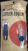 Front of sealed packaging for Chef Works Urban Apron, Galveston bib apron shown in dark blue with adjustable neck strap and front pockets.