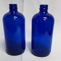 Front view of two 16oz blue glass lotion bottles standing upright on a white background, showing their smooth glossy blue surface and threaded necks.
