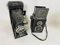 Side by side view of Kodak folding bellows camera and Voightlander Brillant box camera on white background.