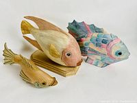 Three carved wooden fish shown, two natural wood finish and one painted pastel colors, displayed on white background