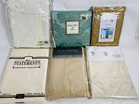 Six tablecloth sets in original packaging, various colors and patterns including lace and solid colors.