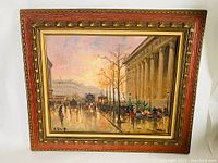 Frontal photo of the Parisian street scene oil painting showing the cityscape with columns, people, and horse carriages under a cloudy sky, in a decorative wooden frame with gold and red detailing.