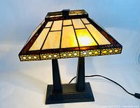 Front view of stained glass desk lamp showing illuminated cream and amber glass panels with geometric borders and metal base with decorative patterns.