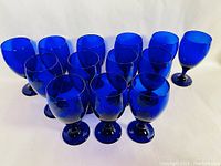 Thirteen cobalt blue glass goblets arranged in a group on a white surface, showing the full lot and color tone.