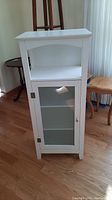 Front view of tall white cabinet with glass door and open shelf above it.