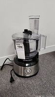 Full view of the Hamilton Beach food processor showing stainless steel and black body with clear plastic lid and chute.