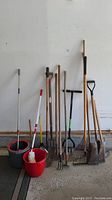 Nine long-handled tools and two mop systems against a white wall