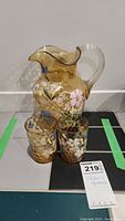 Full view of amber glass pitcher with clear handle and two matching hand painted glasses, all featuring floral designs.