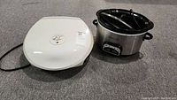 Photo shows white George Foreman grill and silver Crock Pot slow cooker with black ceramic insert and glass lid side by side on carpeted floor.