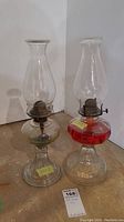 Photo of two clear glass hurricane oil lamps with glass chimneys. One has a clear round base, the other has a red ribbed base. Both have metal burners.
